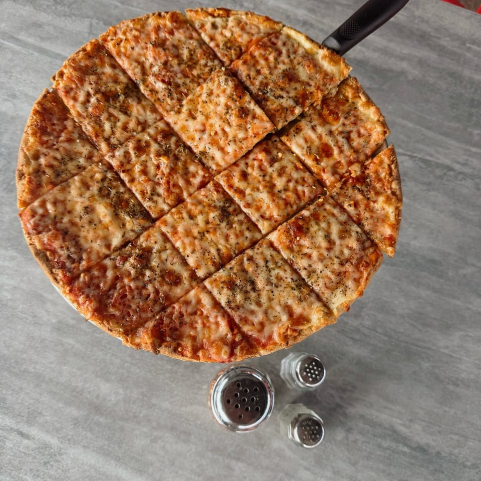 Round cheese pizza, cut into squares, with a server and shakers on a gray table.