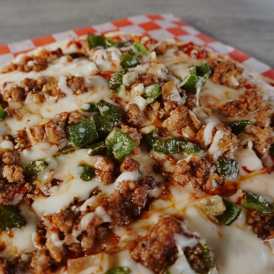 Close-up of a pizza with melted cheese, green peppers, and ground meat on a checkered paper.