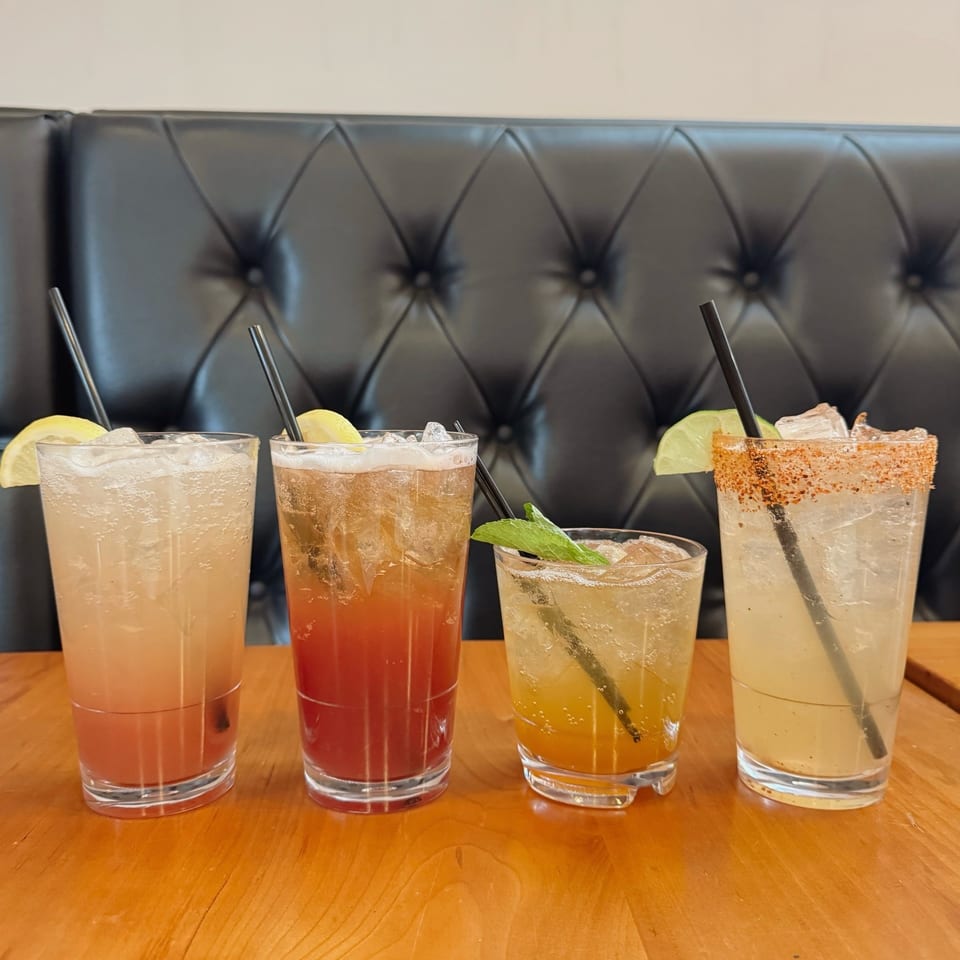 Four colorful cocktails with ice, garnishes, and straws on a wooden table with a black booth.