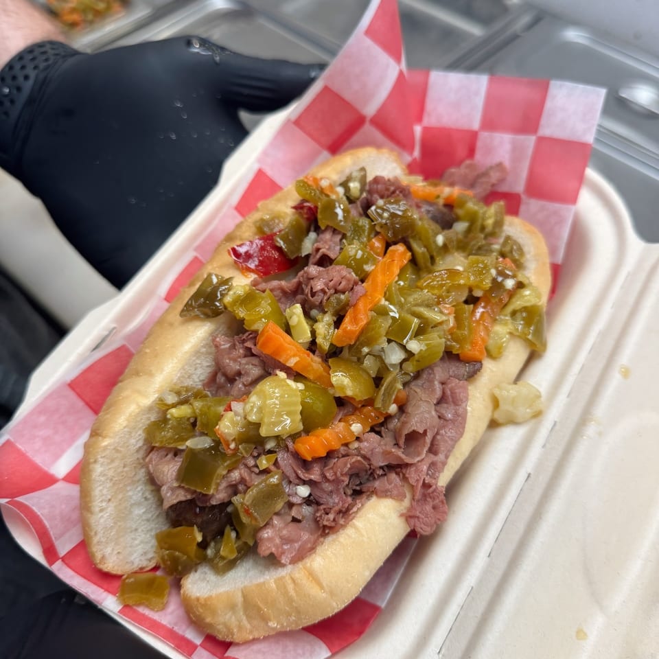 A gloved hand holds a Chicago-style Italian beef sandwich with giardiniera in a white container.
