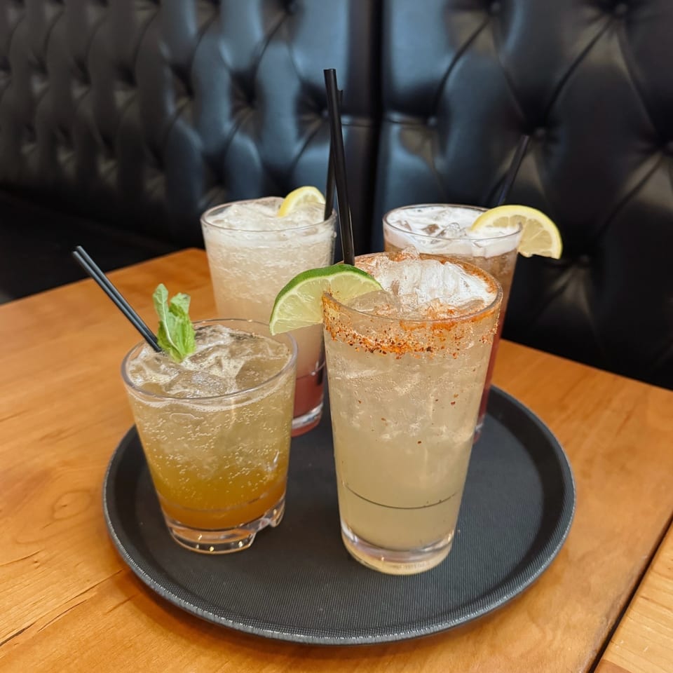 Four colorful cocktails with garnishes on a black tray, on a wooden table with a black booth.